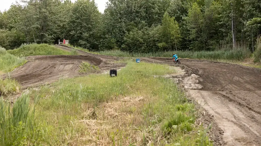 Une personne conduit une moto tout-terrain sur une piste de motocross sinueuse et boueuse, entourée d'herbe verte et d'arbres denses, sous un ciel nuageux.
