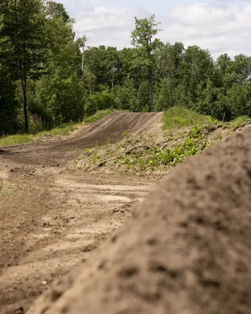 Sous un ciel partiellement nuageux, une piste de motocross serpente à travers une zone herbeuse et bordée d'arbres, avec en arrière-plan des monticules de terre et un feuillage verdoyant.