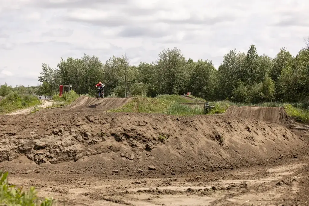Une personne conduit une moto tout-terrain sur une piste de motocross entourée d'arbres et d'un ciel nuageux, avec un terrain accidenté et poussiéreux au premier plan.