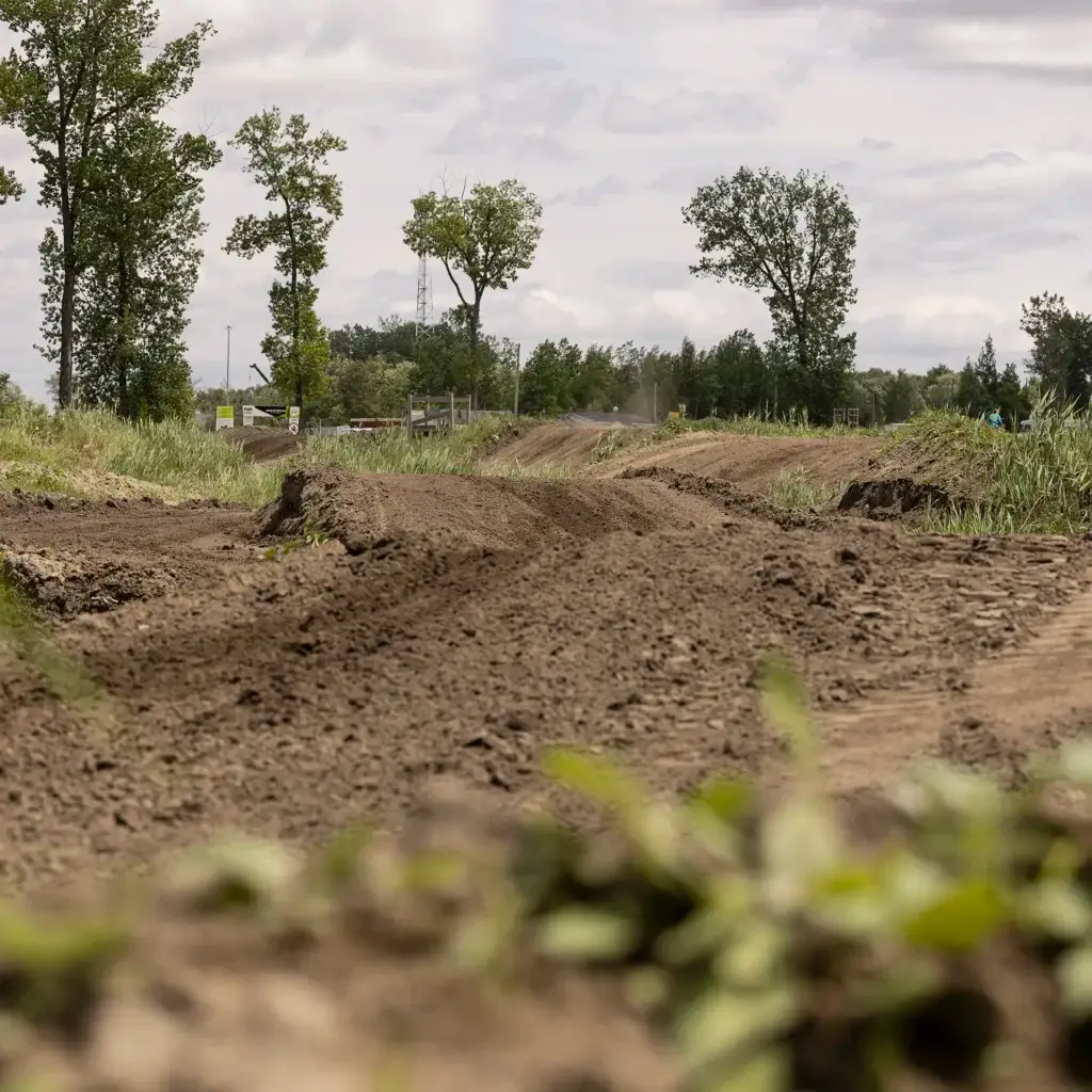 Vue en contre-plongée d'une piste de motocross en terre battue, traversant de l'herbe verte et des arbres sous un ciel nuageux. La surface de la piste est rugueuse et irrégulière, avec de la verdure au premier plan et des arbres à l'arrière-plan.