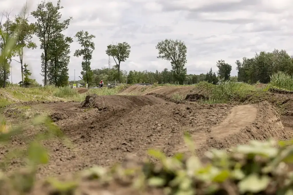 Une piste de motocross en terre battue avec un terrain accidenté et de petits sauts serpente à travers des champs herbeux, entourée de grands arbres sous un ciel nuageux.