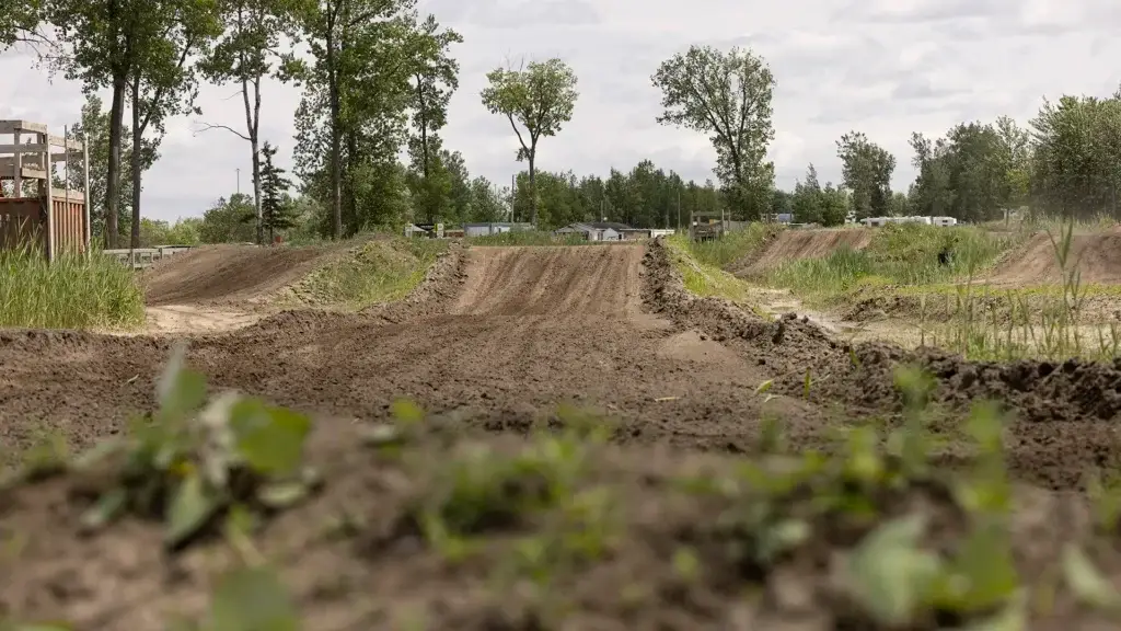 Une piste de motocross en terre battue, avec plusieurs sauts et tremplins, serpente à travers une zone herbeuse et bordée d'arbres, sous un ciel nuageux. La photo est prise en contre-plongée, près du sol.