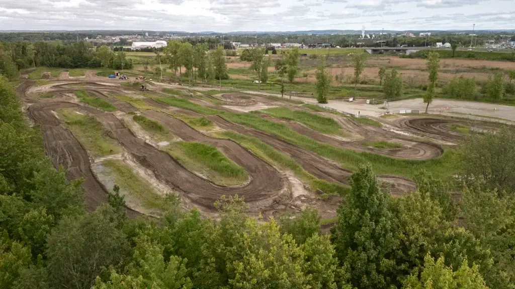 Vue aérienne d'une piste de motocross sinueuse entourée d'arbres verts et de zones herbeuses, avec un ciel nuageux et des bâtiments éloignés en arrière-plan.