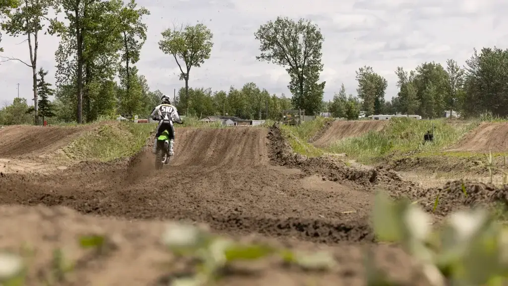 Un pilote de motocross sur une moto tout-terrain court sur une piste accidentée et boueuse, entourée d'herbe et d'arbres, sous un ciel nuageux. Le pilote porte un maillot blanc avec le numéro 501 visible dans le dos.