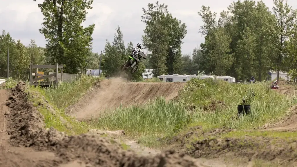 Un pilote de motocross vêtu d'un équipement fait sauter une moto tout-terrain par-dessus un monticule sur une piste extérieure herbeuse, en soulevant de la poussière. Des arbres et plusieurs véhicules récréatifs sont visibles à l'arrière-plan sous un ciel nuageux.