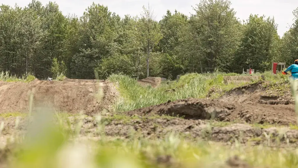 Une piste cyclable traverse un paysage herbeux et accidenté, entouré d'arbres verts et denses. Deux personnes sont visibles : l'une fait du vélo au loin, l'autre se tient à droite, vêtue d'une chemise bleue.