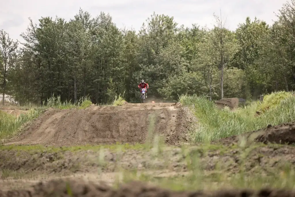 Une personne portant une chemise rouge conduit un vélo tout-terrain sur une piste de terre entourée d'arbres verts et d'herbe, avec des sauts et des monticules visibles sur la piste sous un ciel nuageux.
