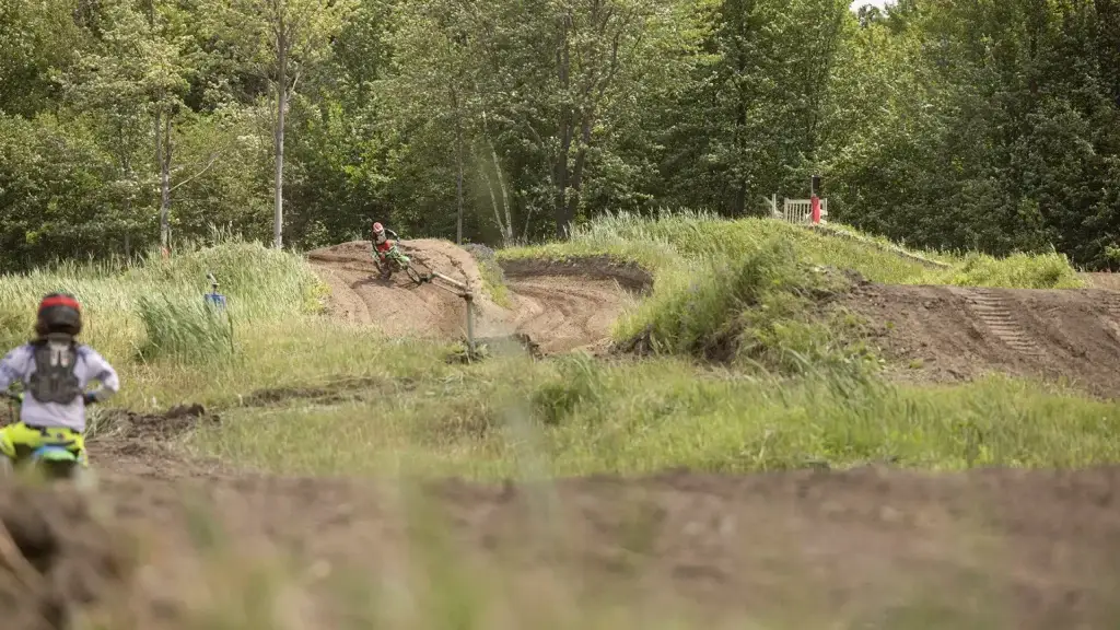 Une personne conduit une moto tout-terrain dans un virage sur une piste entourée d'herbe et d'arbres, tandis qu'une autre personne portant un casque l'observe depuis le premier plan.