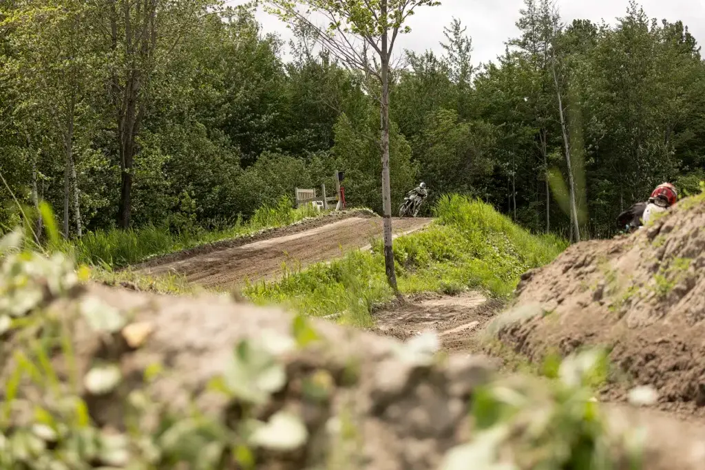 Une personne sur un vélo tout-terrain roule sur un sentier boueux à travers une zone verte et boisée ; une autre personne portant un casque est partiellement visible sur la droite derrière un monticule de terre.