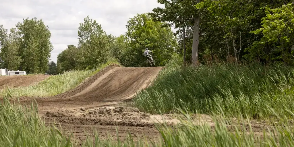 Un pilote de motocross s'envole au-dessus d'un tremplin de piste en terre battue, entouré de hautes herbes vertes et d'arbres par une journée nuageuse.