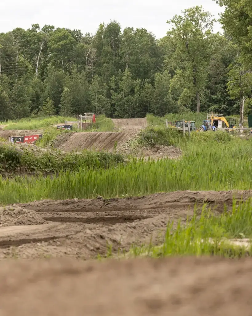 Une piste de motocross avec des traces de pneus serpente dans l'herbe haute, entourée d'arbres verts. Des véhicules et des équipements de construction sont visibles à l'arrière-plan sous un ciel couvert.