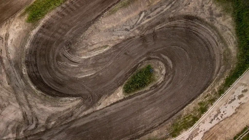 Vue aérienne d'une piste de course en terre boueuse avec des virages sinueux et des traces de pneus. Un petit coin d'herbe se trouve au centre d'un virage, entouré par la piste. La végétation verte borde la piste.