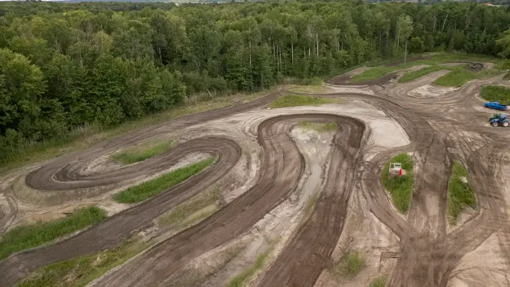 Vue aérienne d'une piste de motocross sinueuse entourée d'arbres verts denses, avec des traces de pneus boueux visibles et un véhicule bleu garé près du bord de la piste.