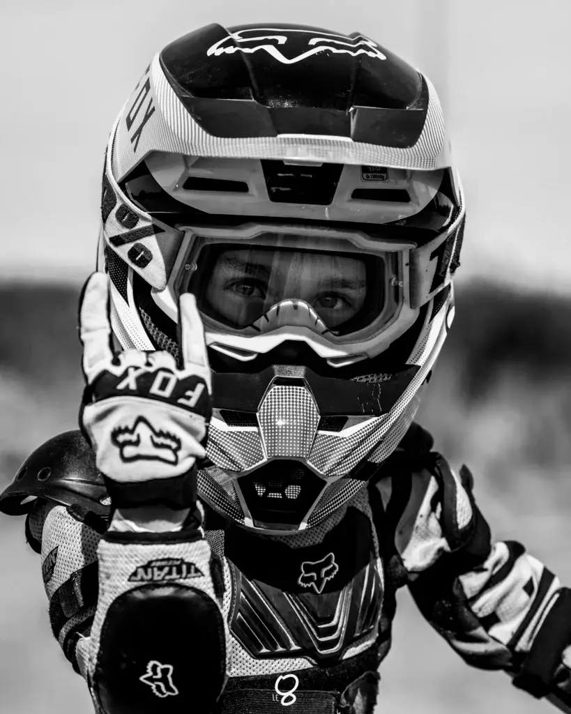 Un pilote de motocross en tenue et casque complet regarde la caméra en levant sa main gantée pour faire le geste "rock on". L'image est en noir et blanc.