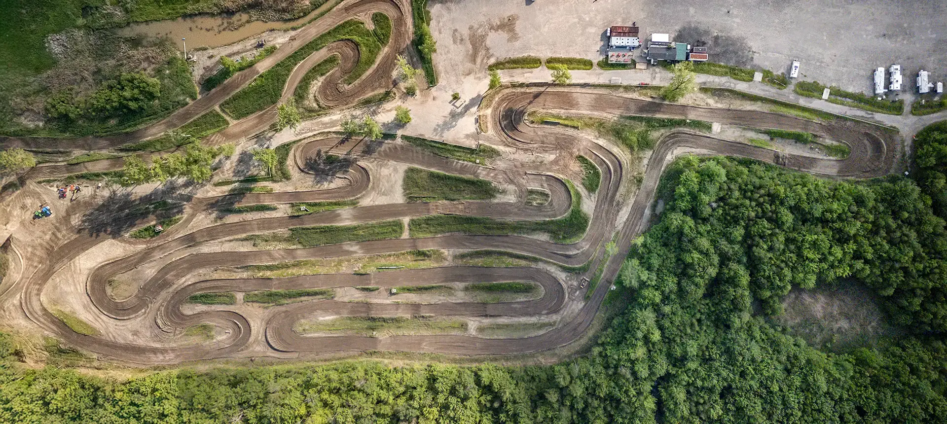 Vue aérienne d'une piste de motocross sinueuse entourée d'arbres, avec une aire de stationnement et plusieurs véhicules près d'un groupe de bâtiments en haut de l'image.