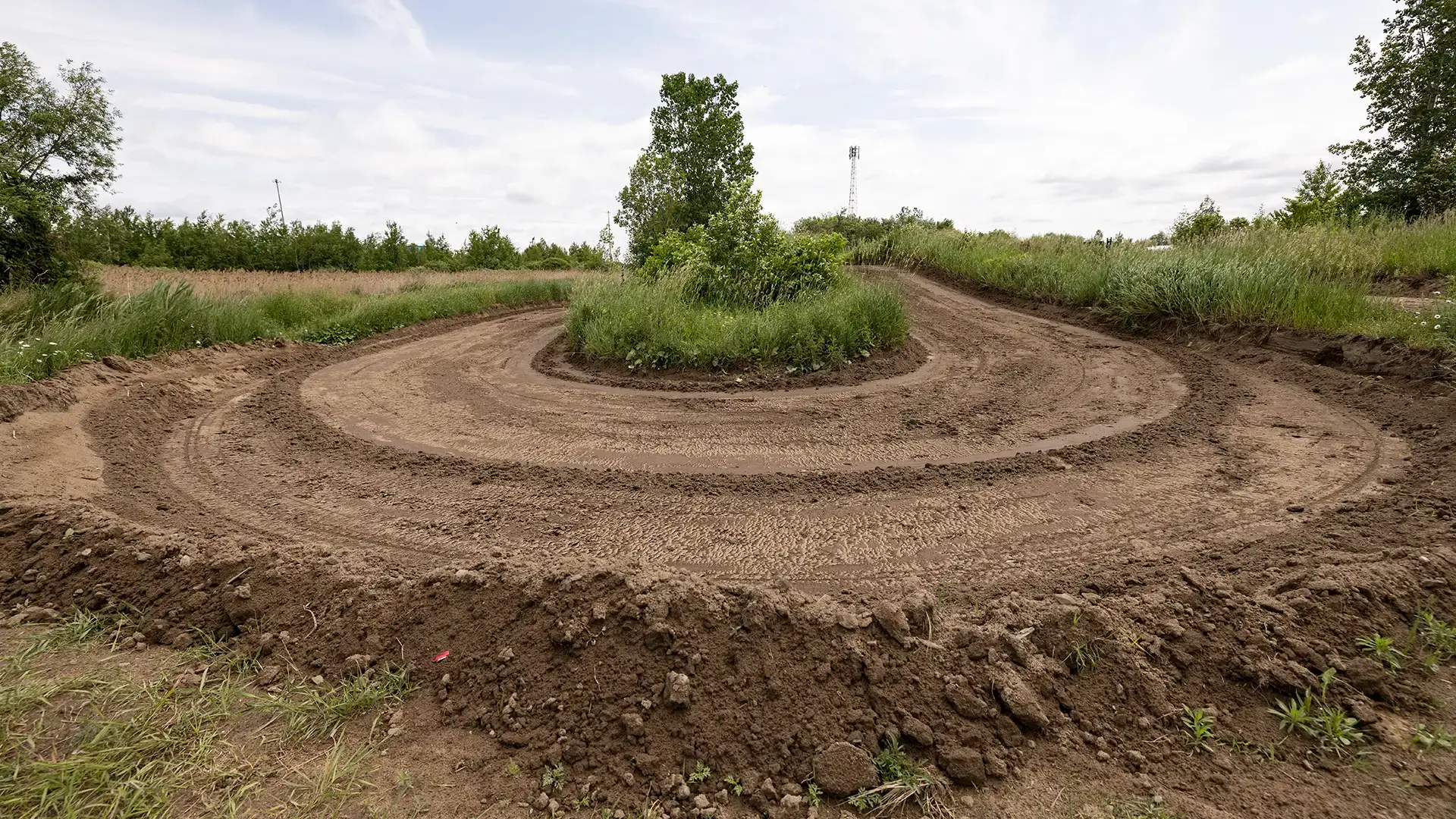 A dirt path winds around a grassy island with trees and tall grass in the center, surrounded by other grasses and a clear sky. The trail appears to have been made recently, with tire tracks visible in the ground.
