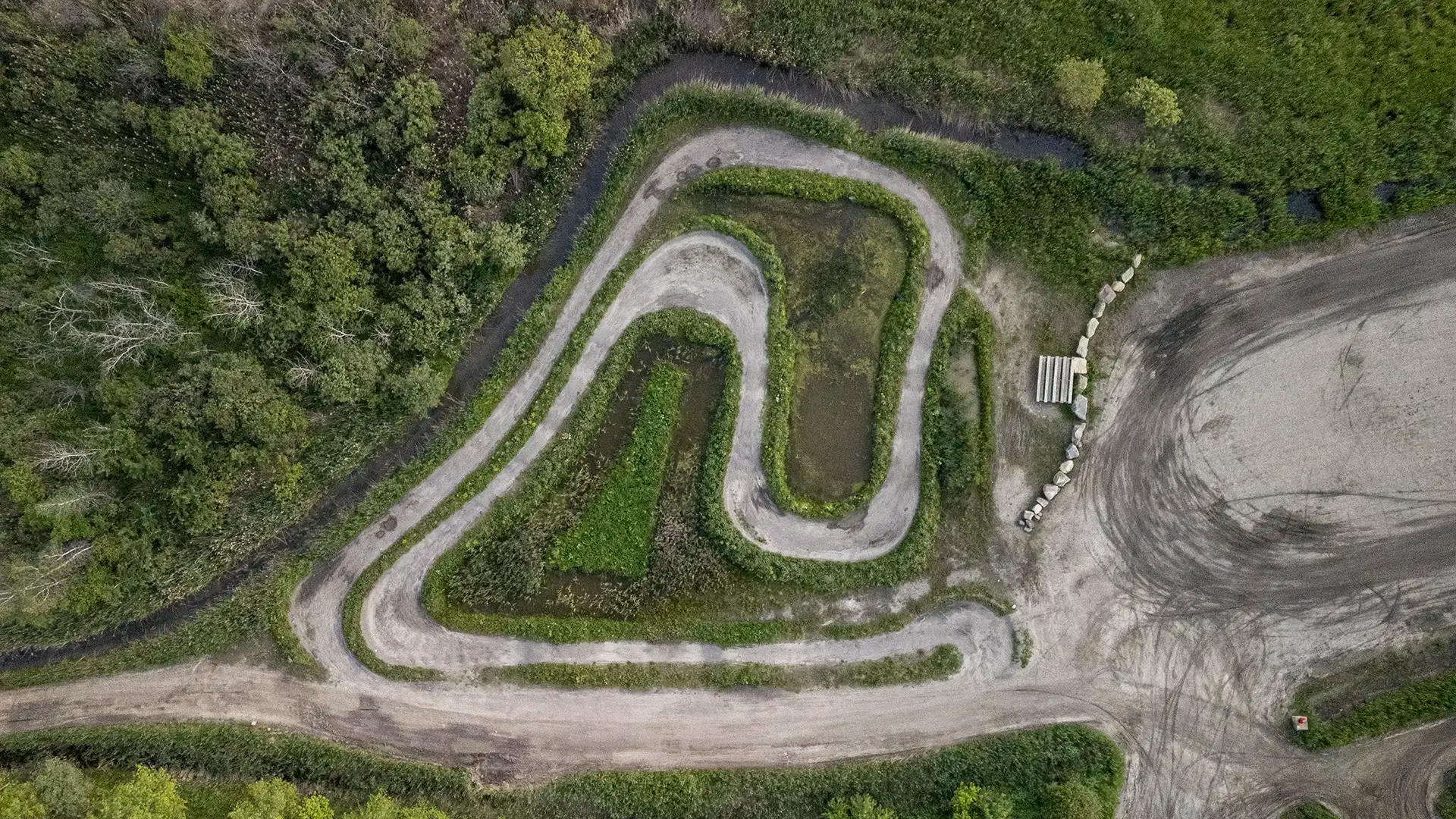 An aerial view of a winding dirt road surrounded by greenery and trees, with a small clearing and scattered rocks on the right side of the image.